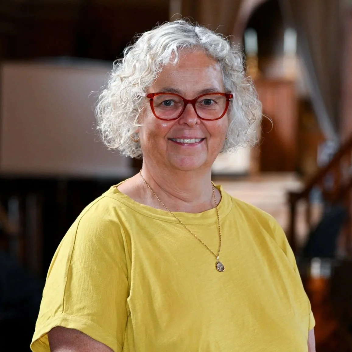 A smiling woman with curly gray hair, wearing red glasses, a yellow shirt, and a gold necklace, standing indoors with wooden decor in the background.