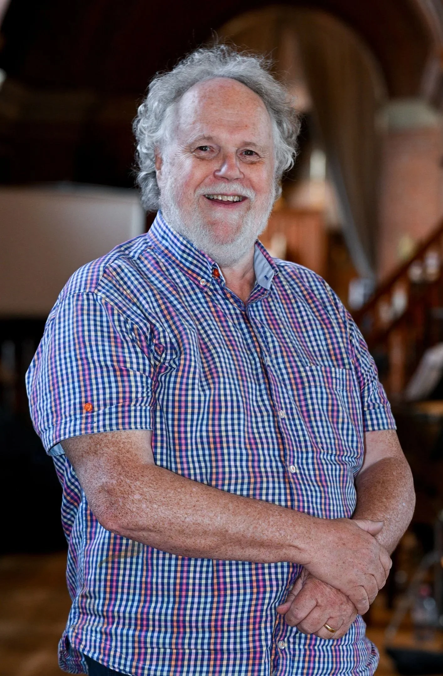 A middle-aged man with gray curly hair and a beard, smiling, wearing a multicolored checkered shirt, standing in a warmly lit room with wooden furniture.