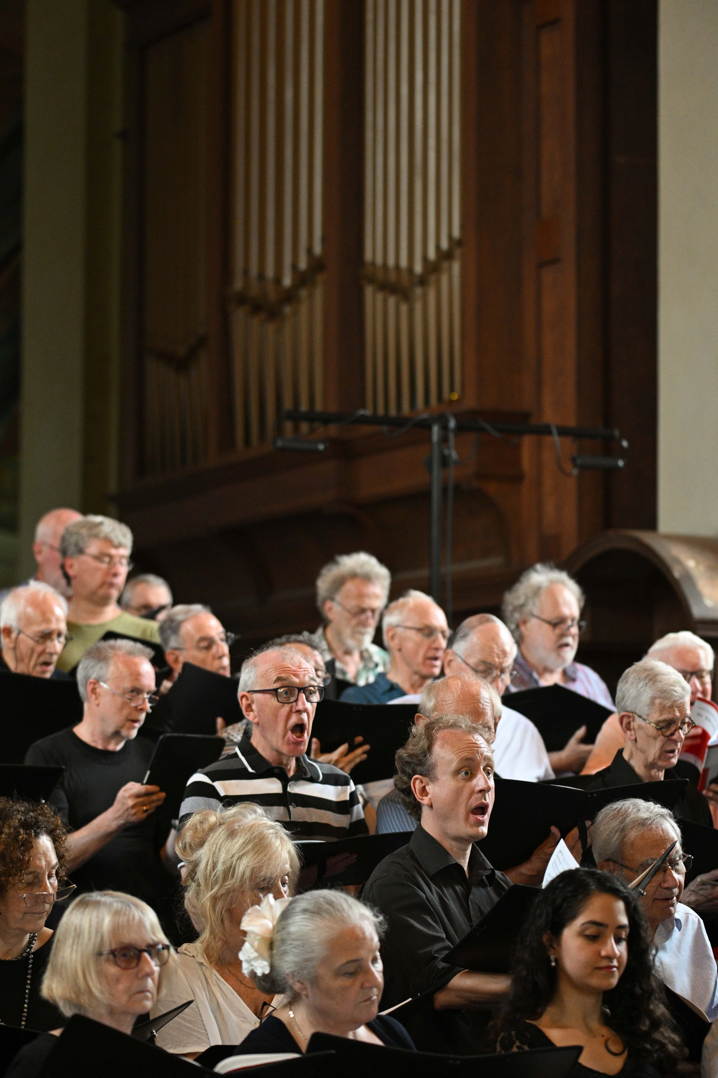 A large group of people singing in a choir, with many holding sheet music, in a church or concert hall. The background features a wooden organ or decorative wood paneling.
