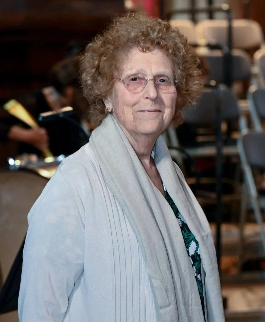 An elderly woman with curly hair and glasses, wearing a light gray jacket, sitting in a auditorium or conference room with chairs and other people in the background.