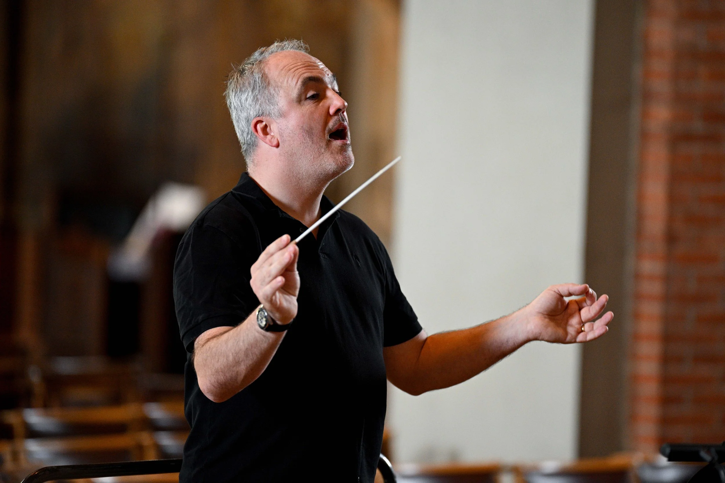 A middle-aged man with gray hair and a beard, wearing a black shirt and a wristwatch, conducting an orchestra or choir with a white baton in his hand in an indoor setting.