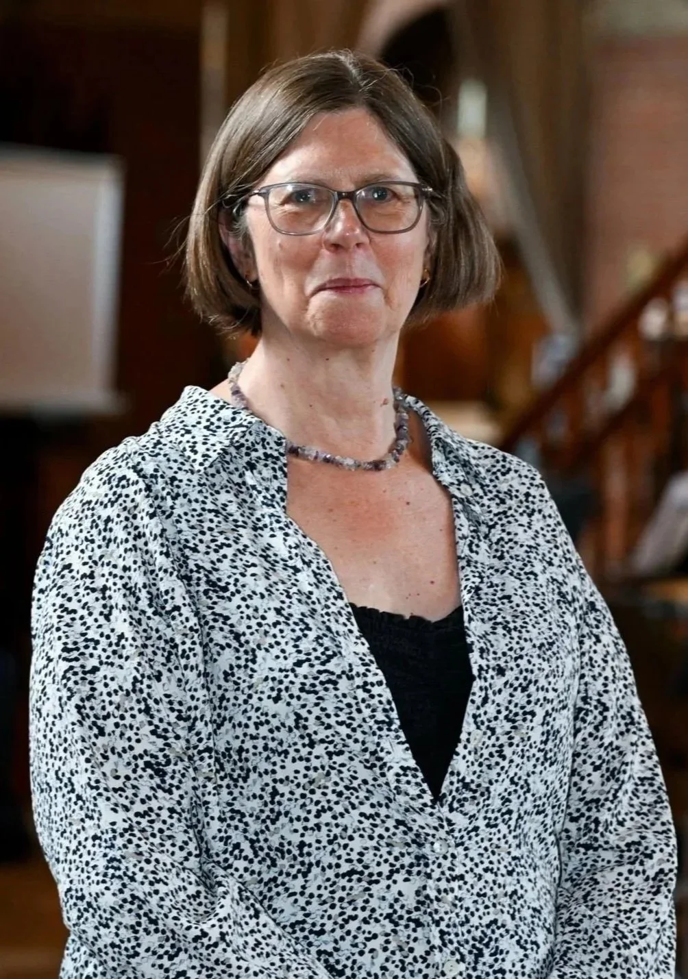 A middle-aged woman with short brown hair, glasses, wearing a patterned blouse, standing in a wooden interior setting.