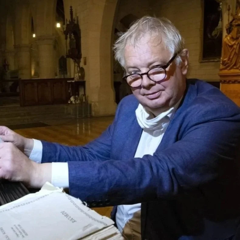 An older man with glasses, curly blonde hair, wearing a blue blazer, white shirt, and a white face mask around his neck, sitting at a desk with papers in front of him inside a church or cathedral with tall arches and religious artwork in the background.