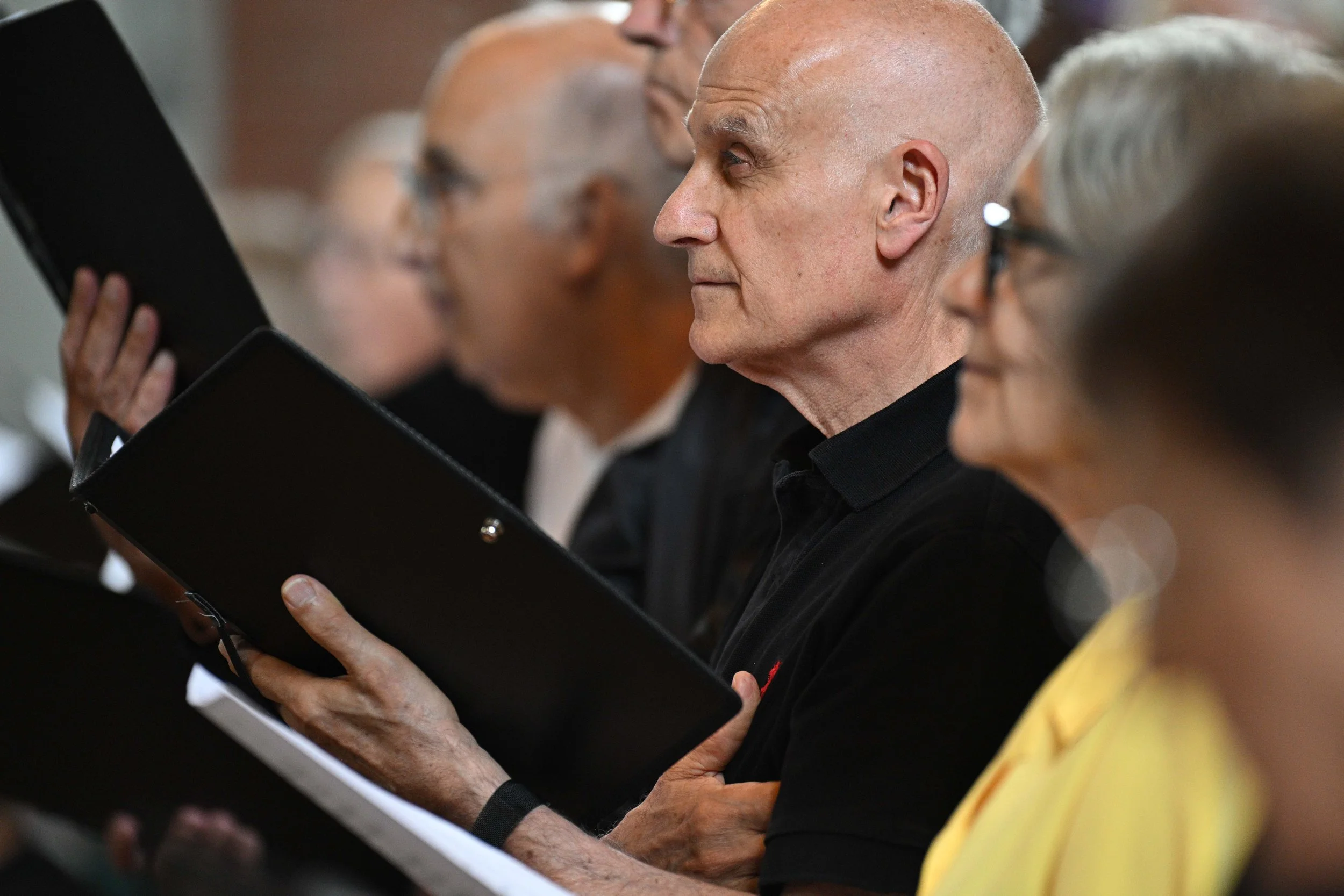 Close-up of a man with a bald head and light skin reading a black folder or binder, surrounded by other people, in a professional setting.