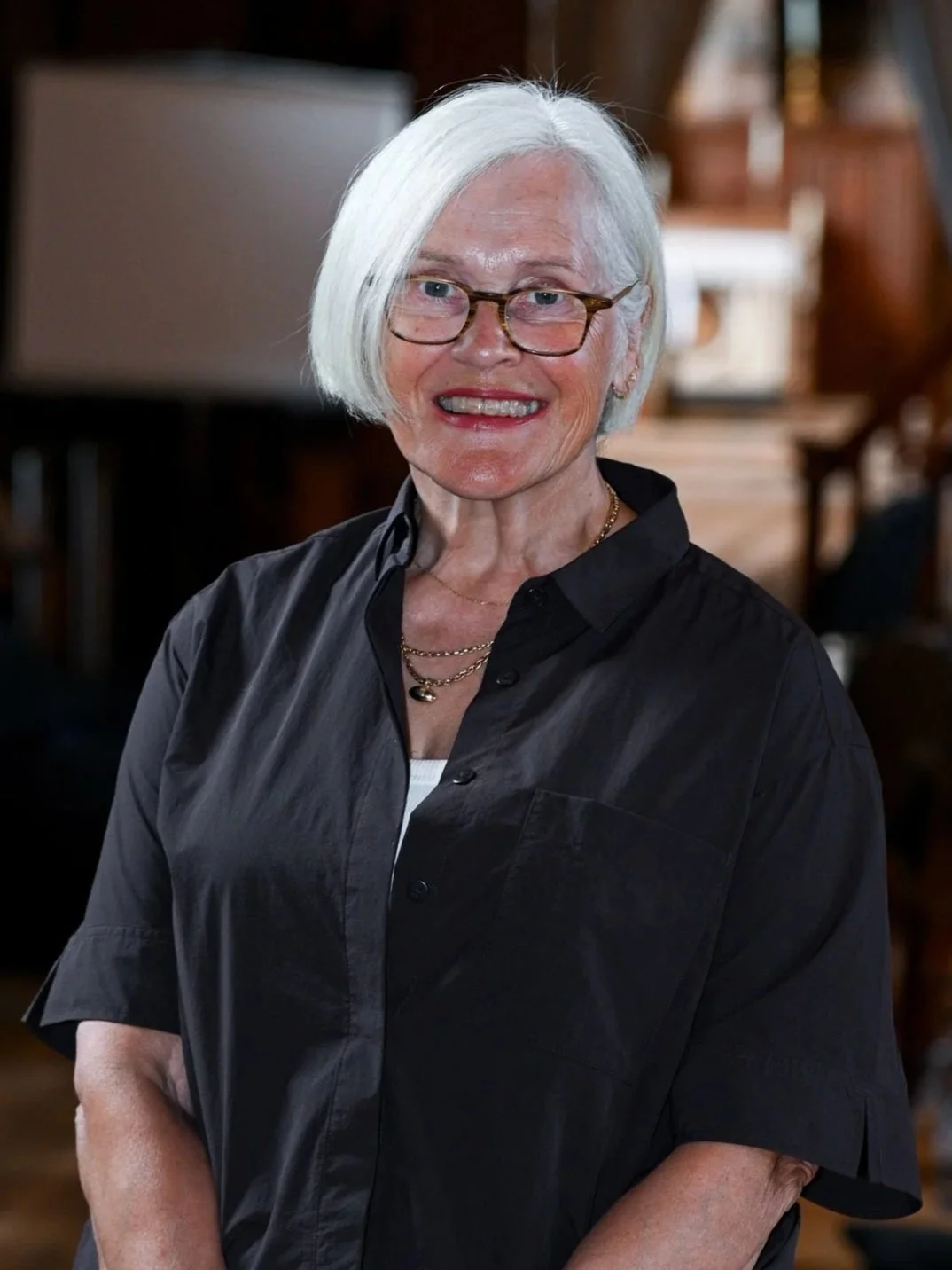 An elderly woman with white hair, glasses, and multiple necklaces smiling in a warmly lit room.