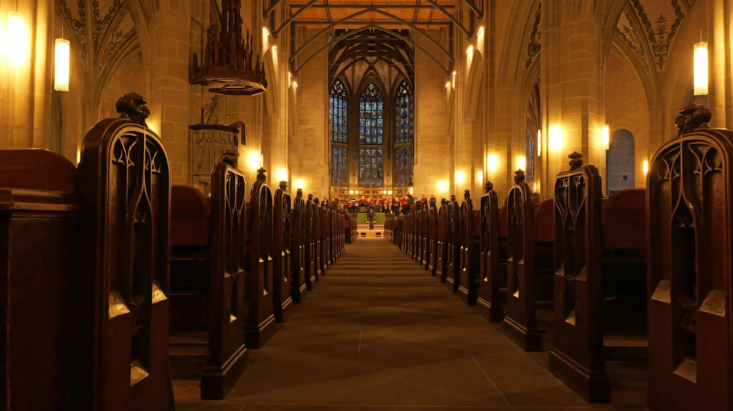 Interior of a church with wooden pews, stained glass windows, and a choir area at the front with people singing.