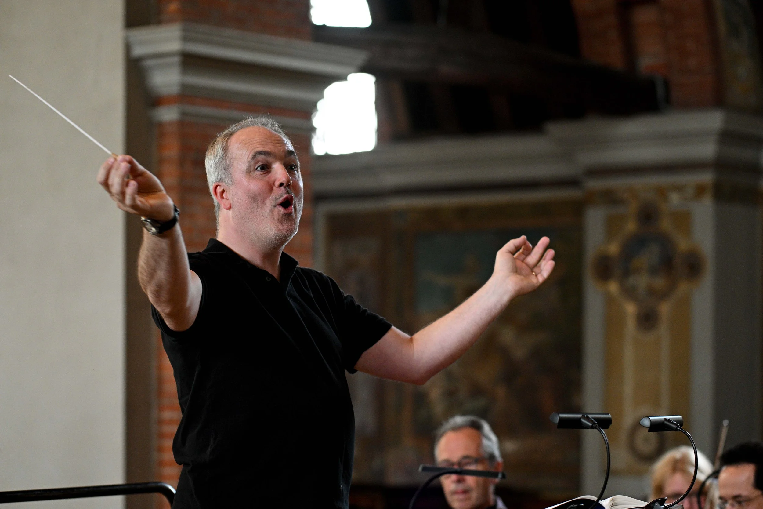 A man with gray hair and a beard, wearing a black shirt, is conducting with a baton in a room with high ceilings and brick walls. Several people are seated in front of him, listening.