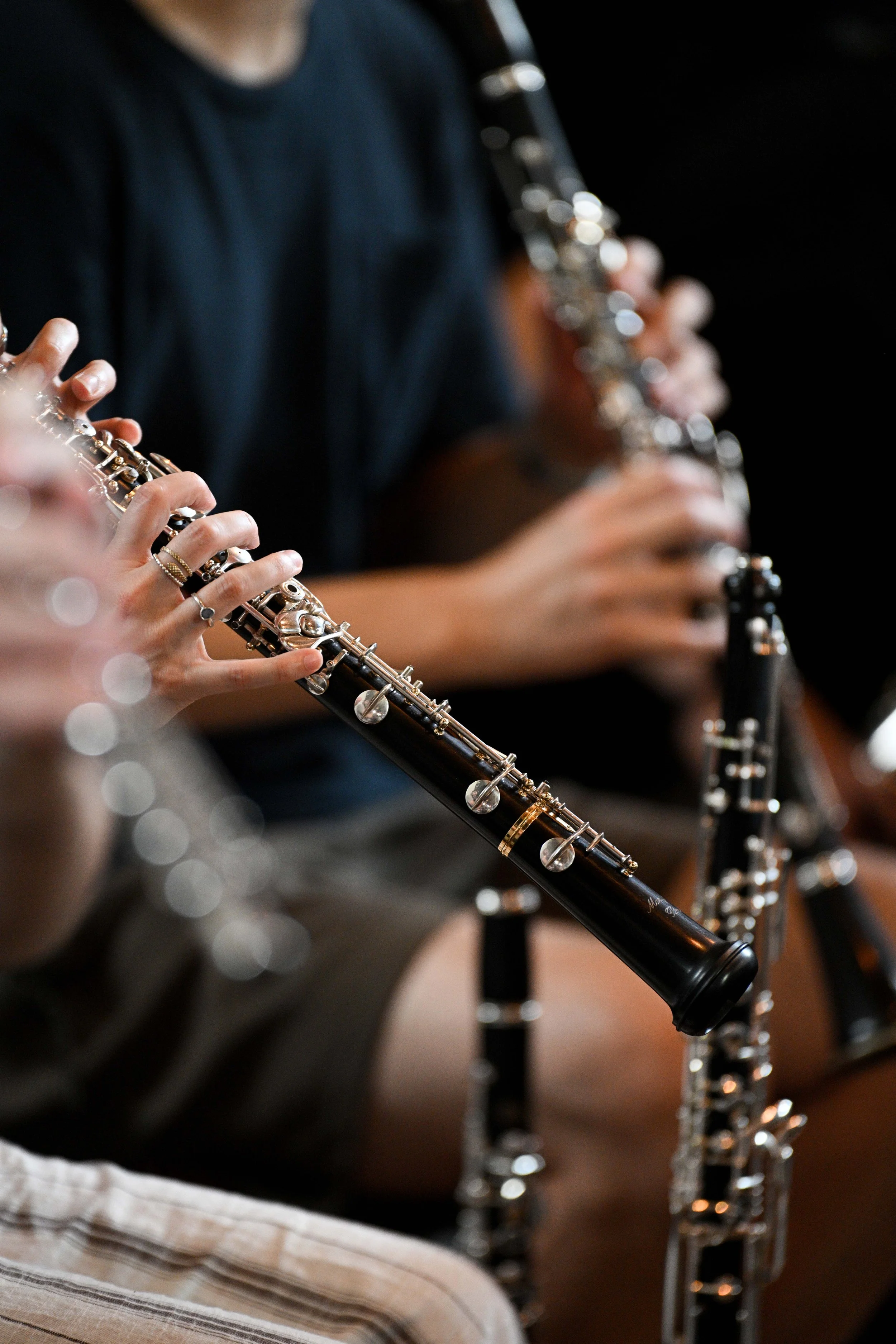 Close-up of a person playing a black and silver flute, holding it with both hands. The person is wearing a dark shirt and is sitting on a chair with other similar instruments in the background. The setting appears to be a performance or practice space.