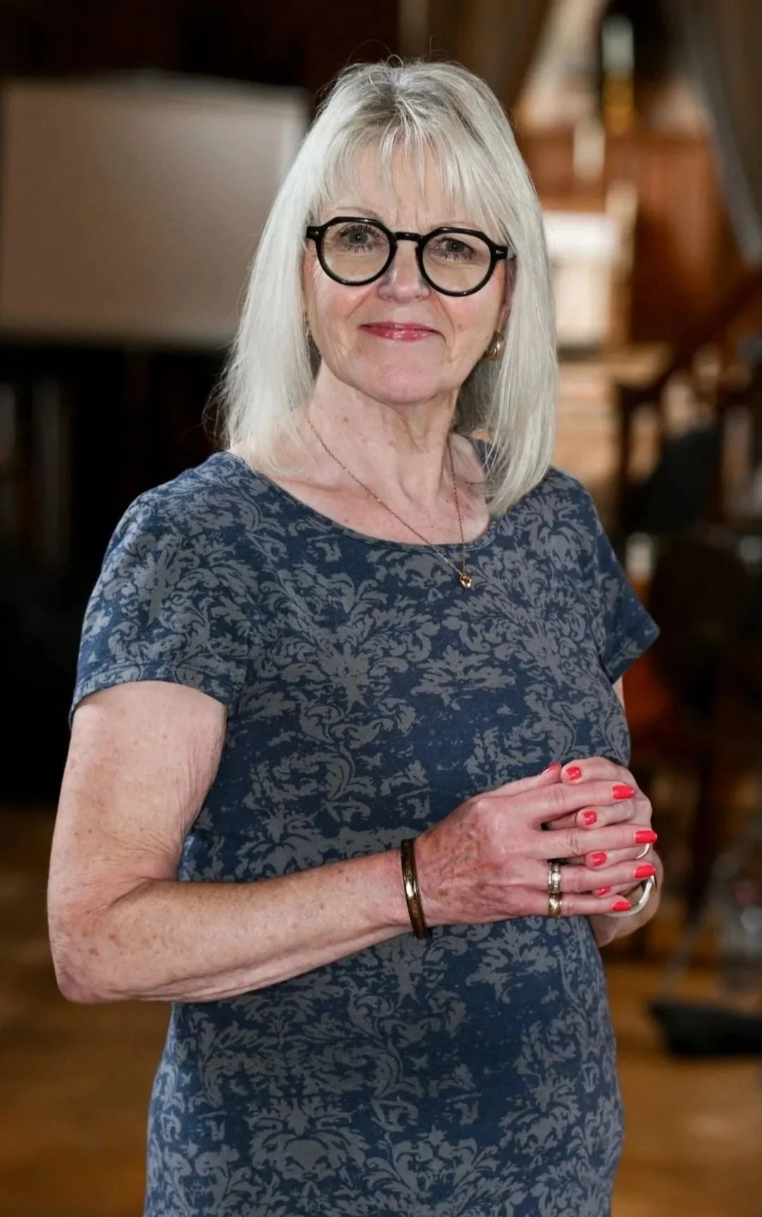 An older woman with blonde hair, glasses, and a floral blue dress standing indoors with her hands clasped.
