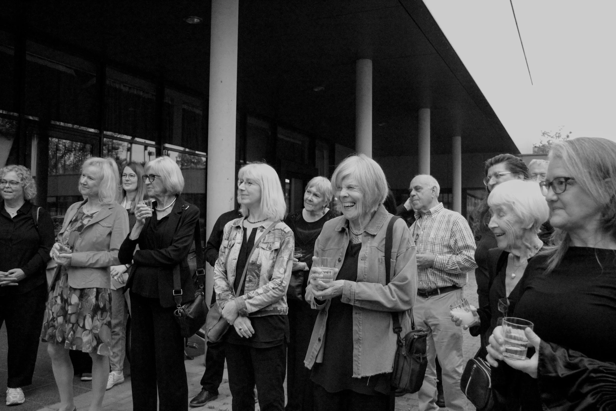 A group of older adults, mostly women, standing outside and smiling at a social gathering, some holding drinks.
