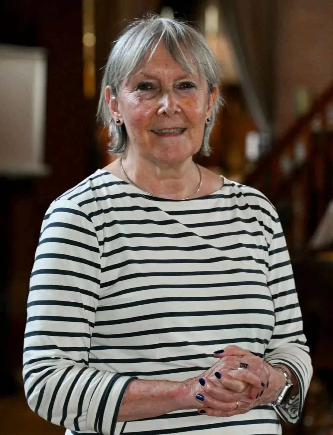 An elderly woman with short gray hair wearing a black and white striped long-sleeve shirt, smiling and clasping her hands together in a warmly lit indoor setting.