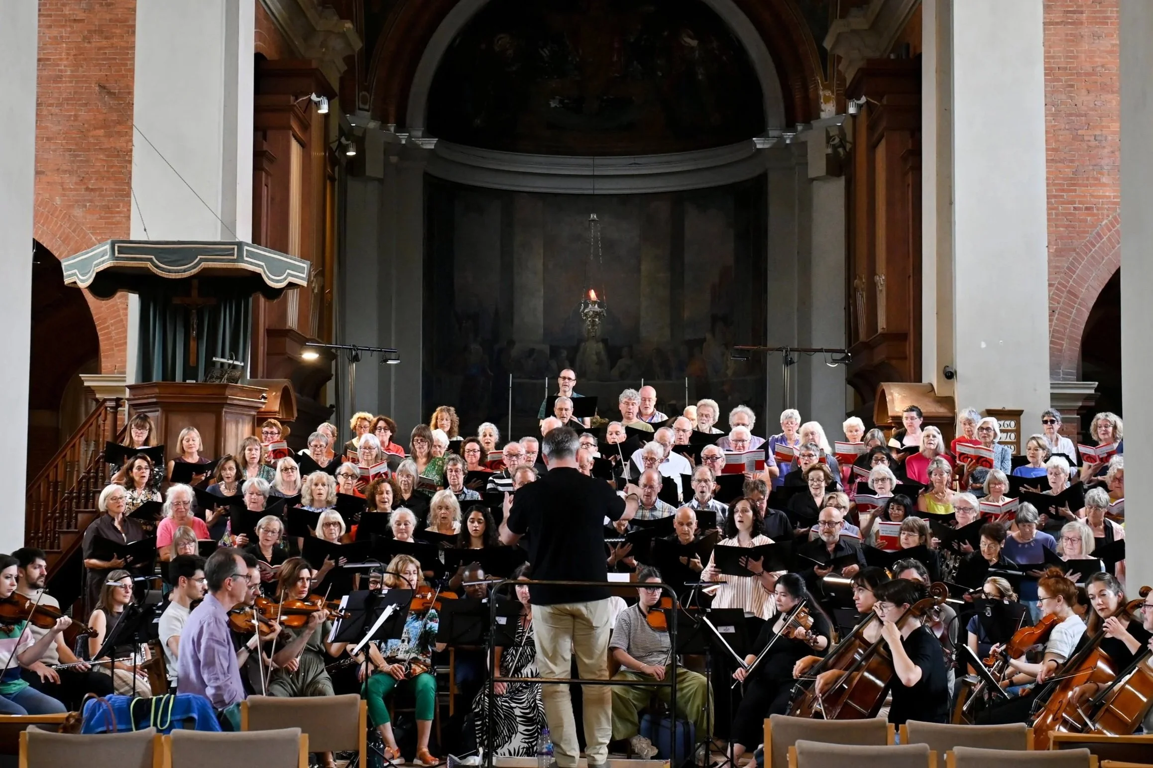 A choir and orchestra performing inside a church with a conductor leading them, several musicians playing violins, and a large group of singers holding sheet music, with church architecture visible in the background.