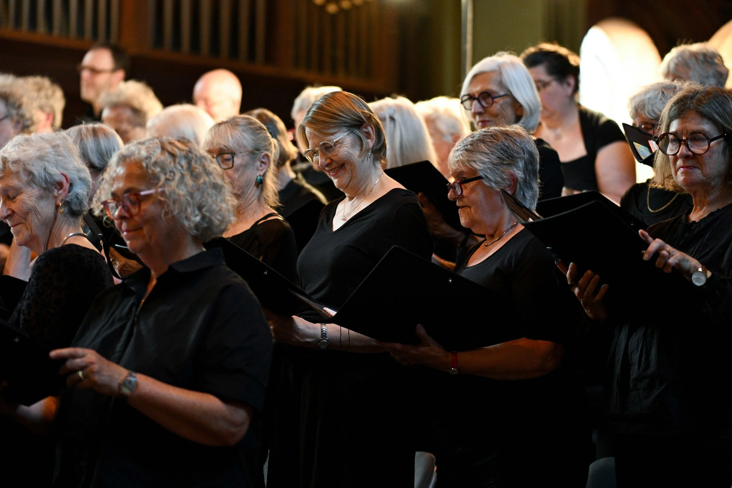 A group of elderly women dressed in black are standing and singing during a choir performance, holding sheet music or folders.
