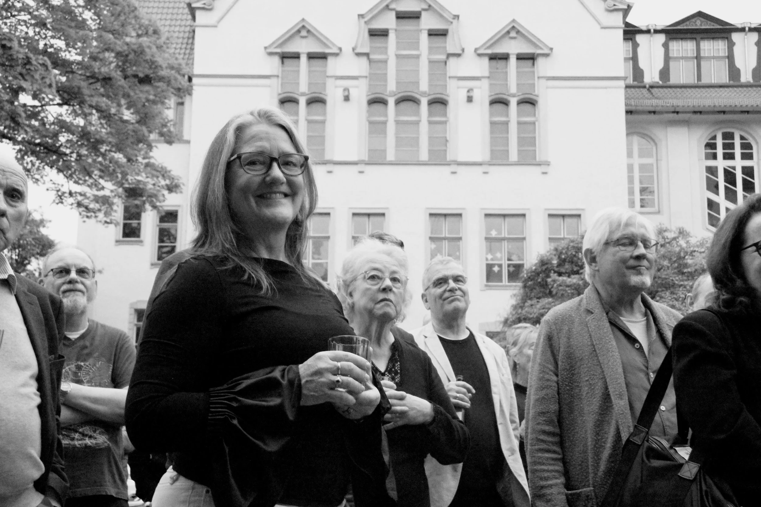 A group of mostly middle-aged and older adults gathered outdoors in front of a large building with ornate windows and a steeply pitched roof. The image shows a smiling woman with glasses in the foreground, holding a drink, with her arms crossed, and 