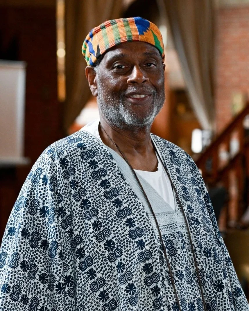 An older man wearing a colorful kente hat and a patterned dashiki, standing indoors with a warm wooden background.