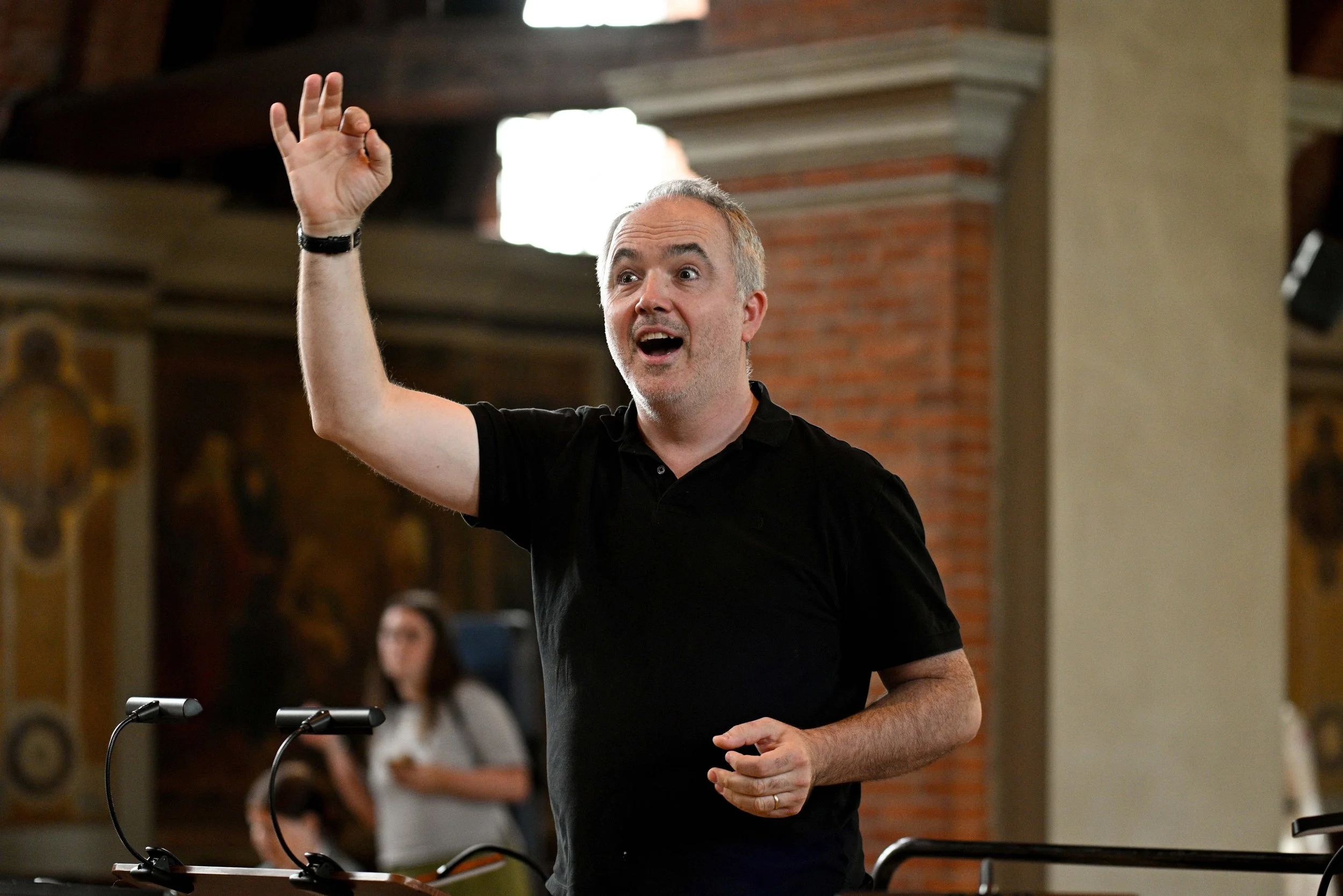 A man with gray hair and a beard, wearing a black shirt, is speaking or giving a presentation in a room with brick and painted walls. He has his right hand raised with fingers forming an OK sign, and appears animated and expressive.