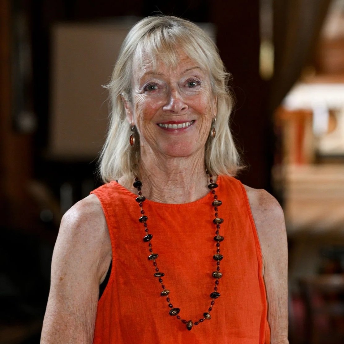 An elderly woman with light blonde hair smiling, wearing an orange sleeveless top and a black beaded necklace, indoors with blurred background.
