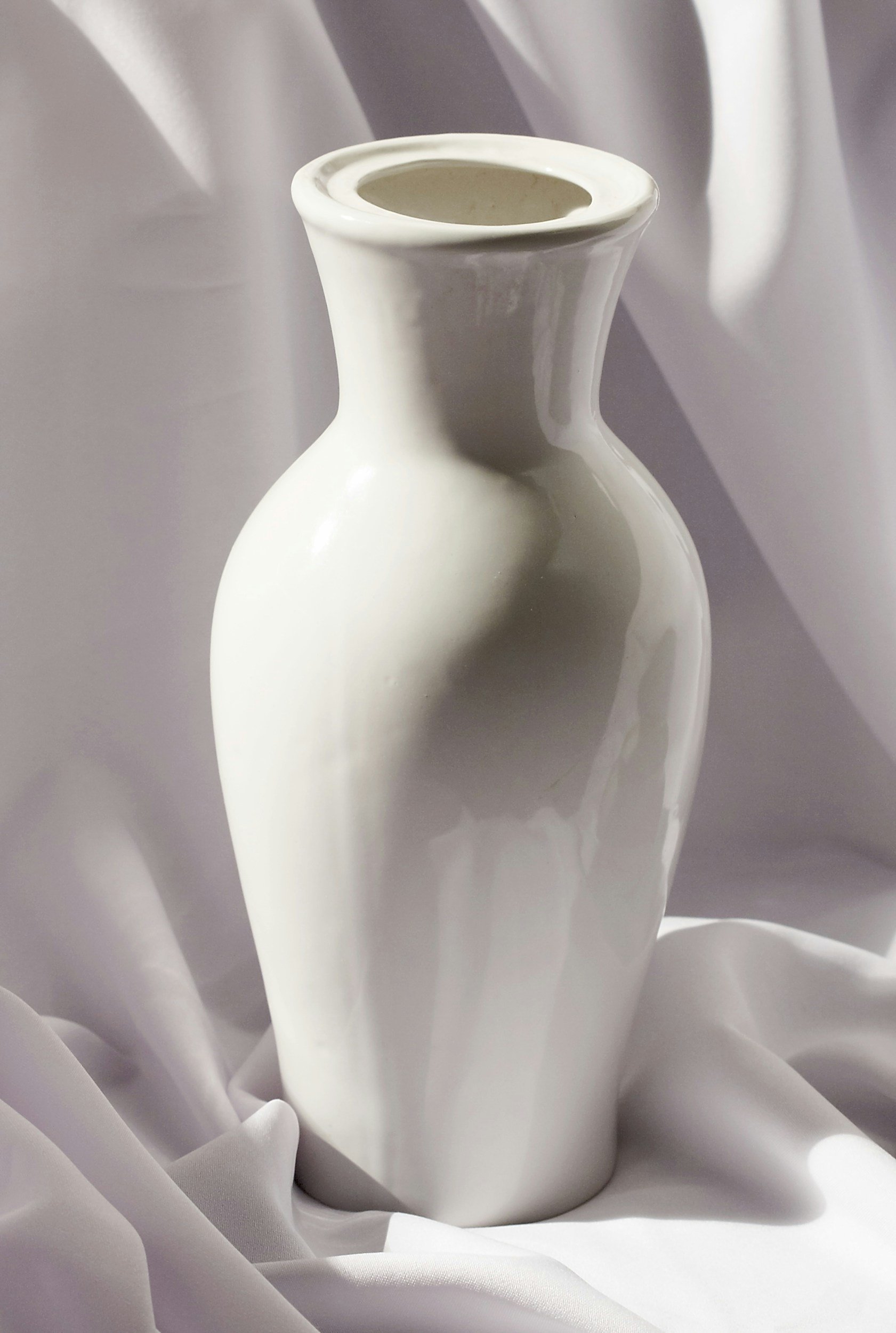 A white ceramic vase with a narrow neck and rounded body, set against a white fabric background with soft folds.