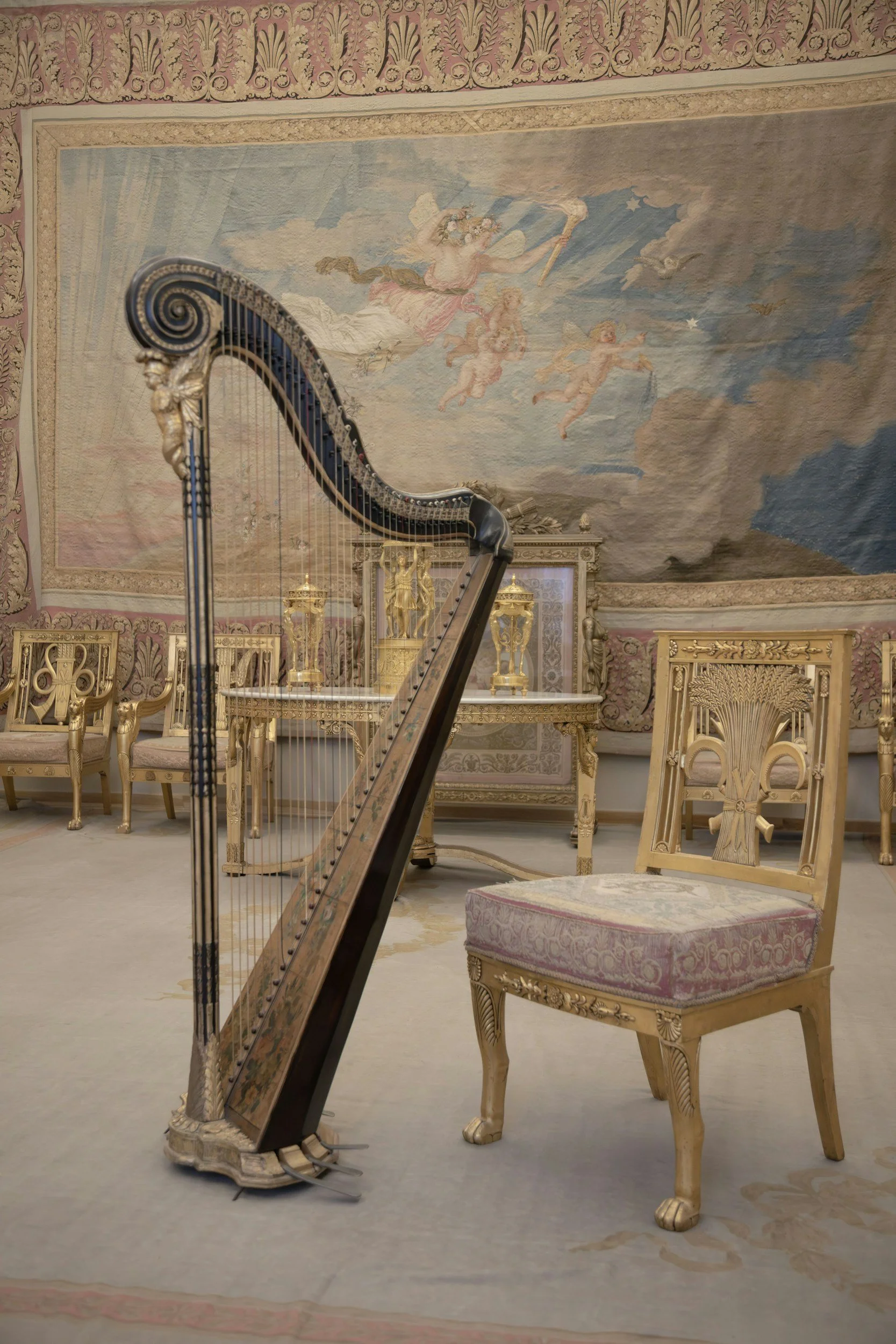 An ornate room with a grand harpsichord, antique chairs, and a large decorative rug on the wall depicting angels, clouds, and a starry sky.