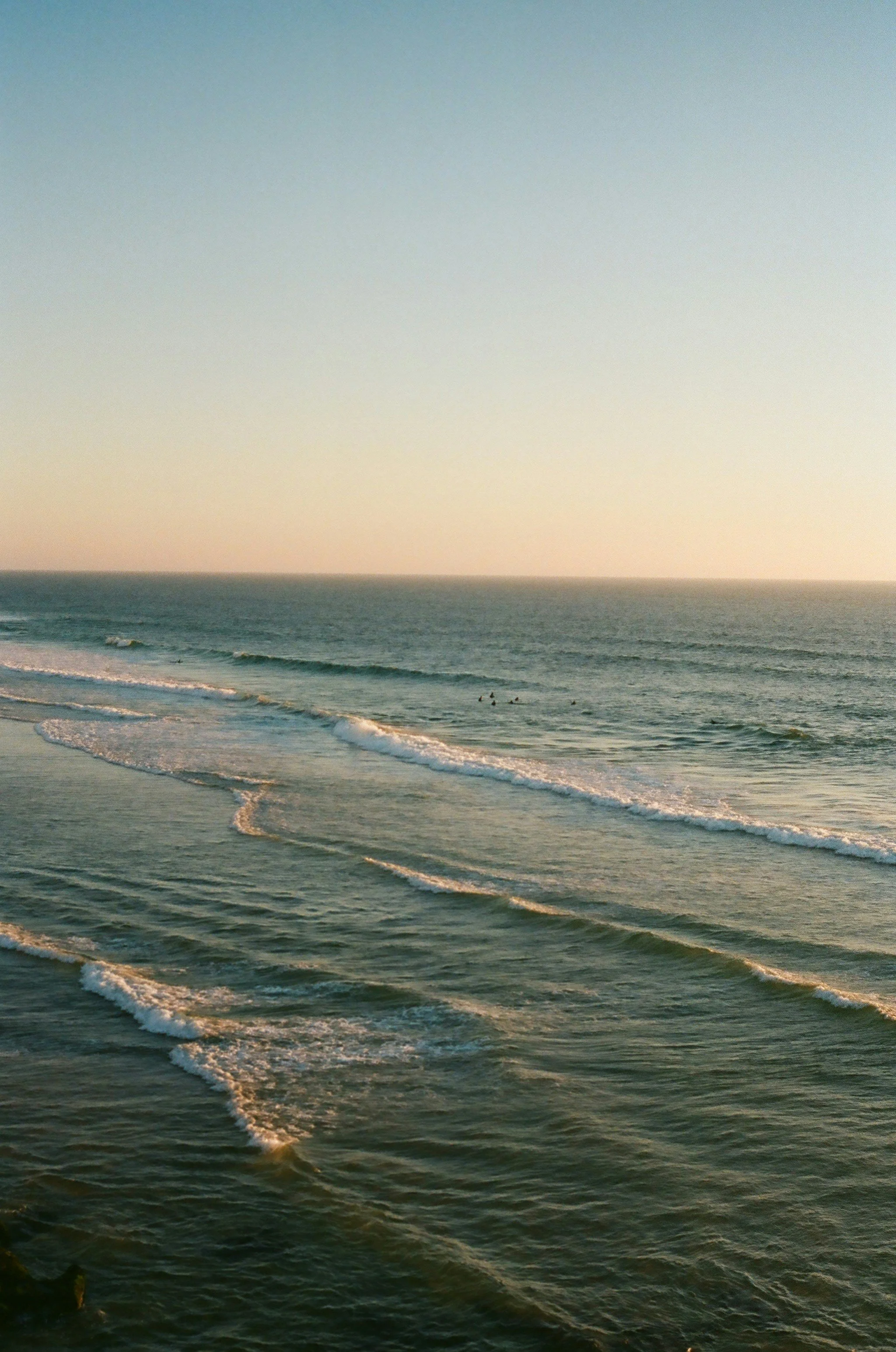 A view of the ocean with gentle waves rolling toward the shore during sunset or sunrise, with a pastel sky in the background.