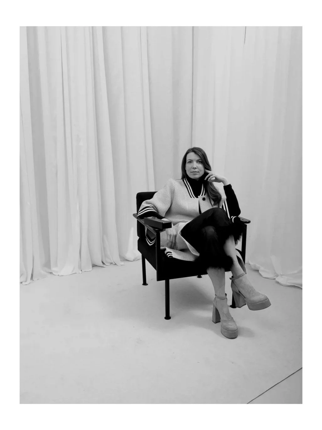 A woman sits on a modern chair with a black seat and back, against a backdrop of plain, swooping curtains, wearing a light-colored coat, black turtleneck, and high heeled boots.