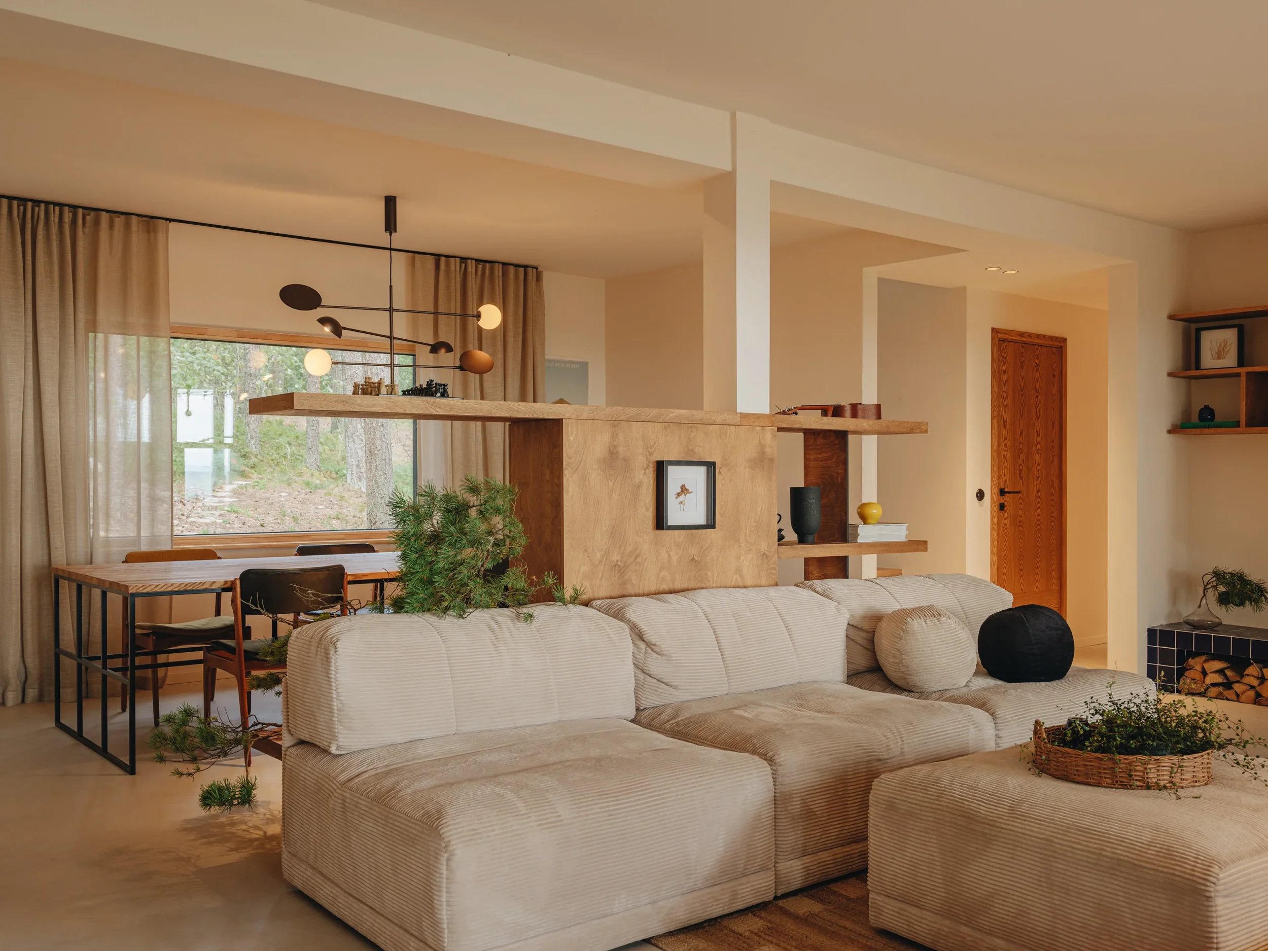 Living room area with beige sofa, wooden dining table, large window with beige curtains, and decorative plants.