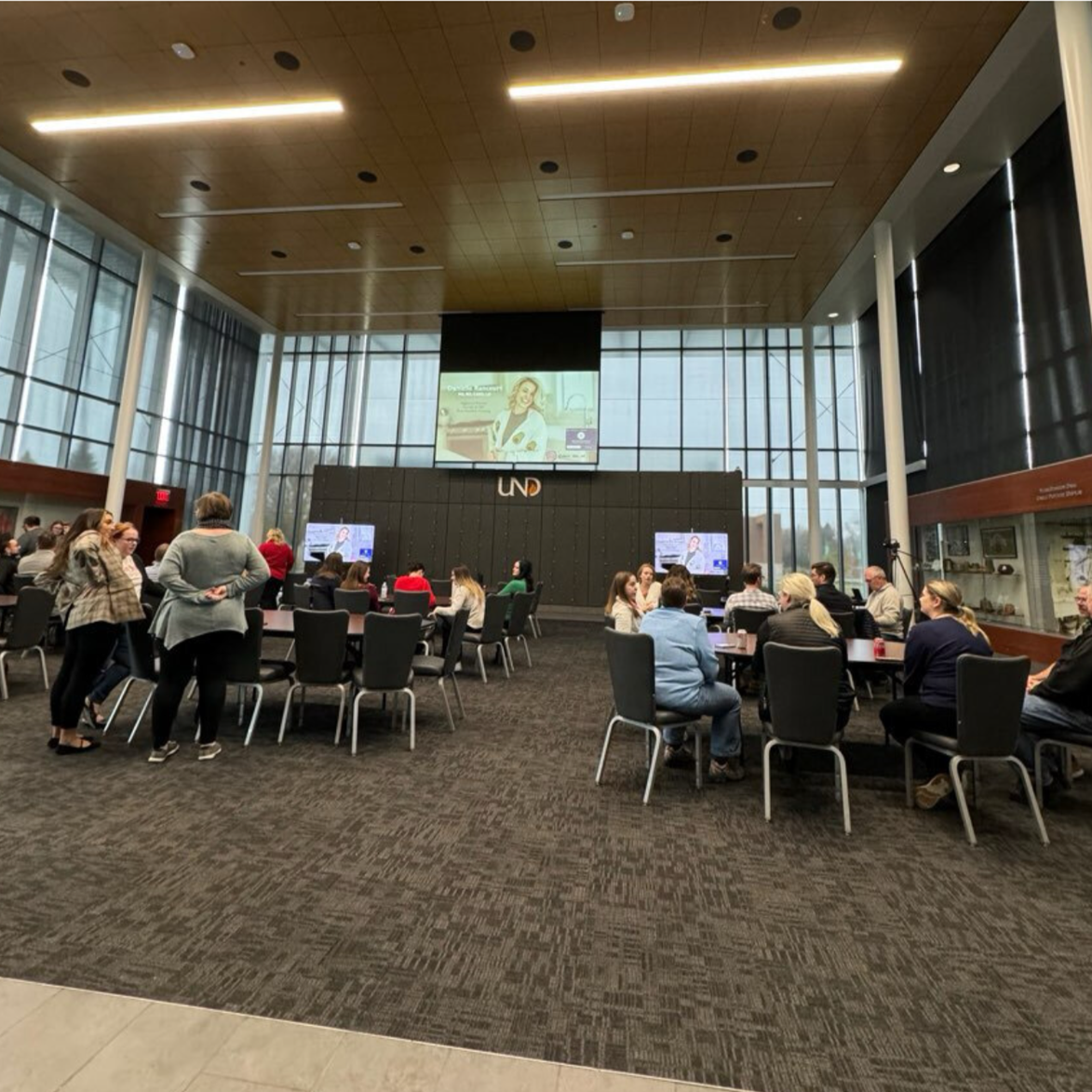 A conference or seminar taking place in a large, modern hall with high glass walls and a wooden ceiling. People are seated at tables, engaging in discussions or listening to a speaker. There are multiple screens, including a large central one displaying a presentation, and the logo 'UND' on the stage backdrop.
