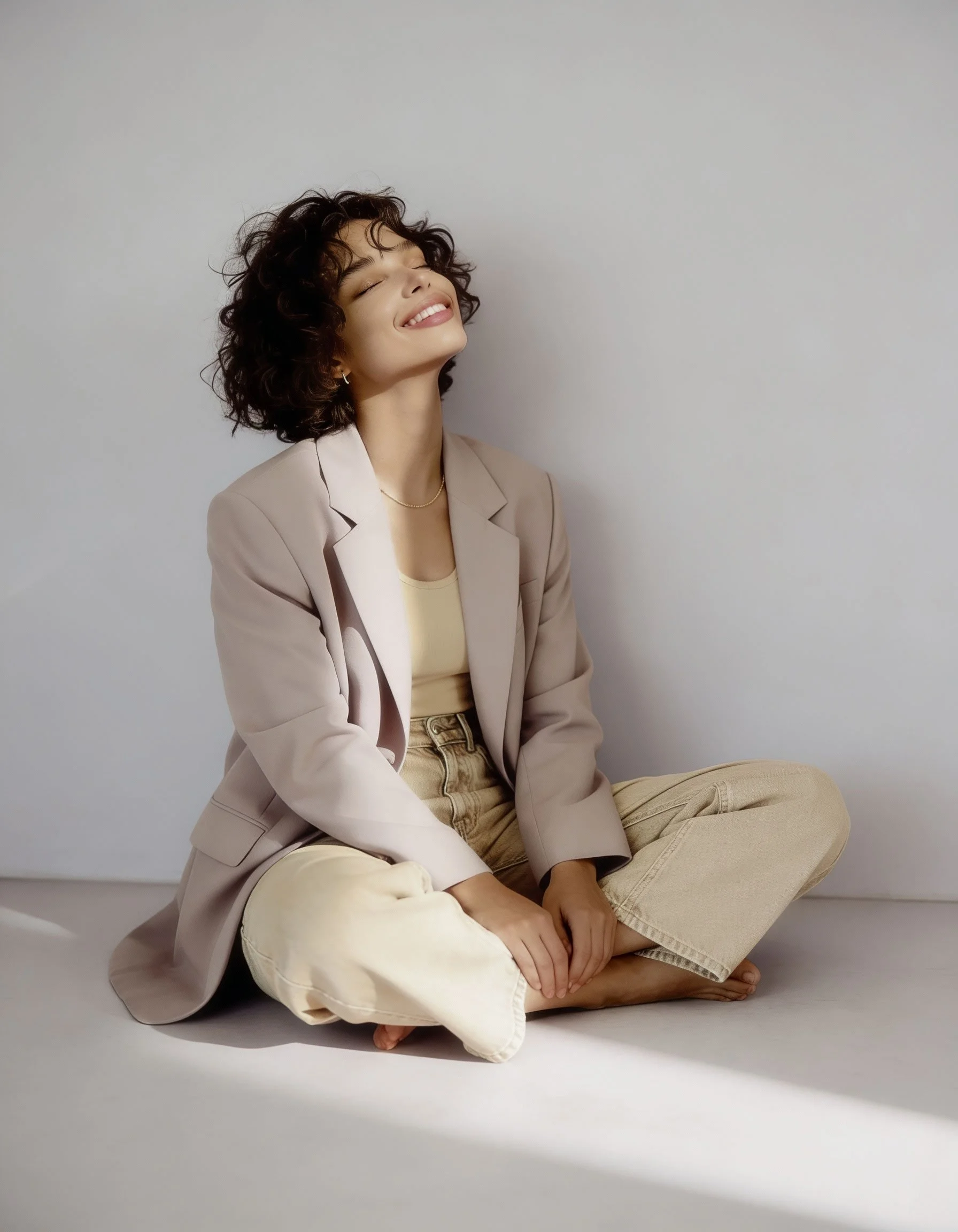 A woman with curly dark hair and light skin sits cross-legged on the floor against a plain white background, smiling with eyes closed, wearing a beige suit jacket and cream-colored pants.