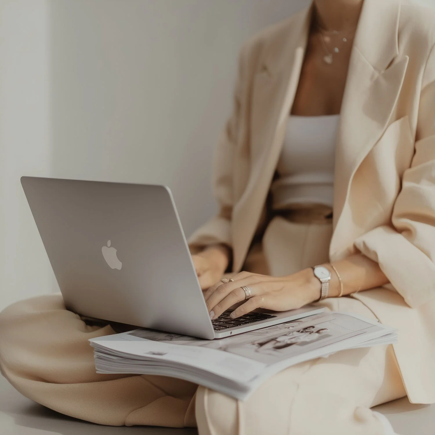 Person wearing a beige suit working on a silver MacBook laptop, sitting on a beige cushion with magazines.