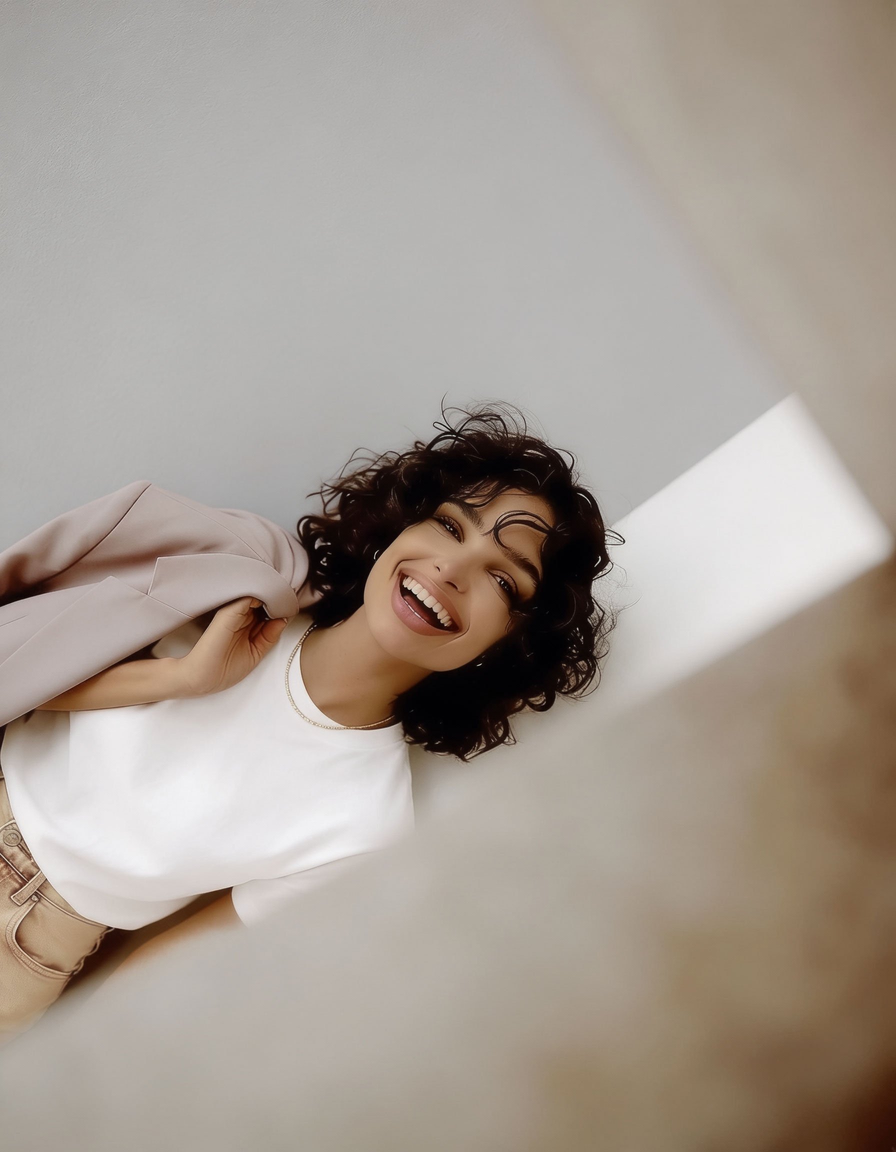 A woman with curly dark hair smiling and looking at the camera, wearing a white top and beige blazer, standing against a plain neutral background.