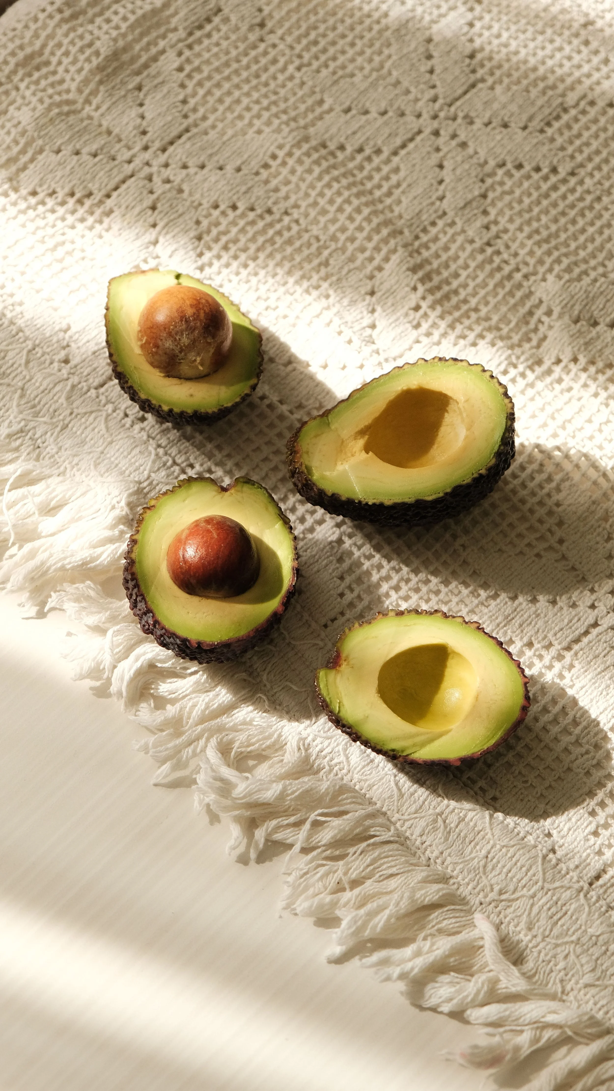Four halved avocados with pits on a textured white cloth with fringed edges, lit by natural sunlight.