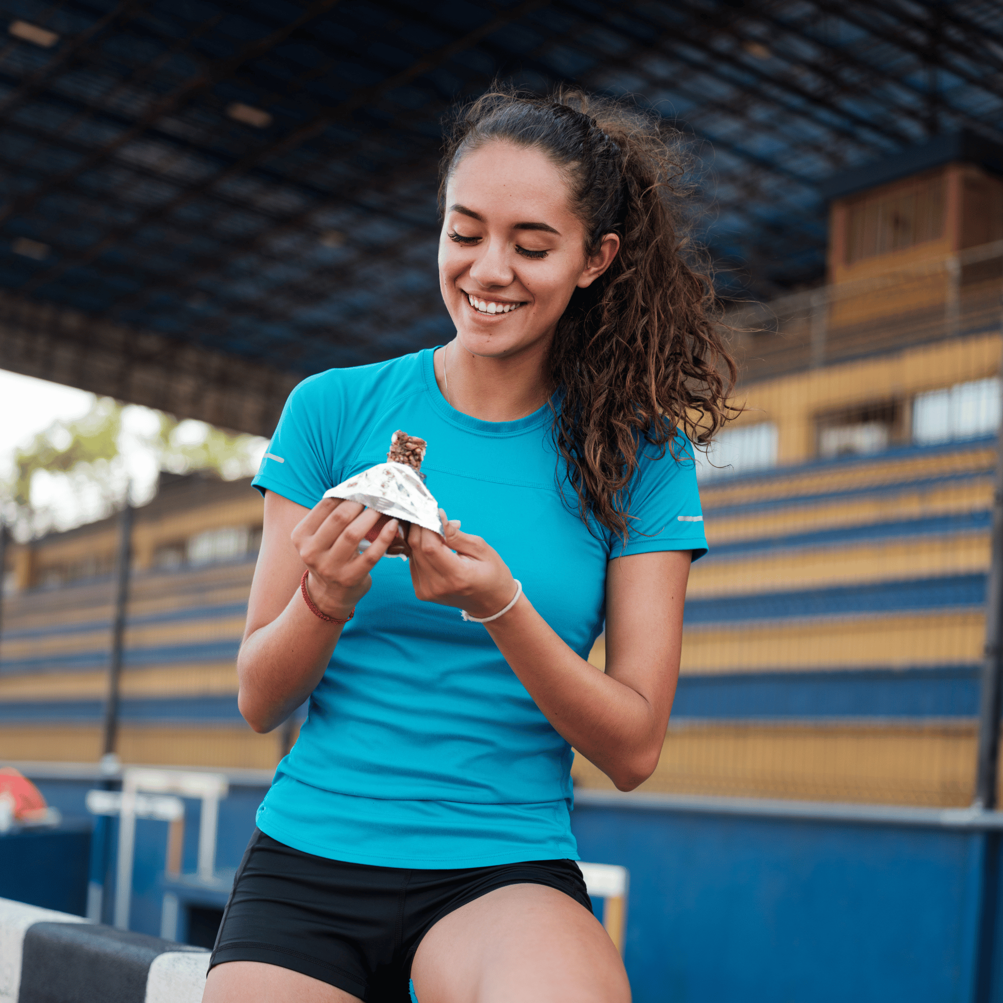 A young woman with curly brown hair wearing a blue athletic shirt and black shorts, smiling as she holds a protein bar wrapped in foil, standing outdoors under a large metal structure. Certified Sports Dietitian approach to fueling athletes.