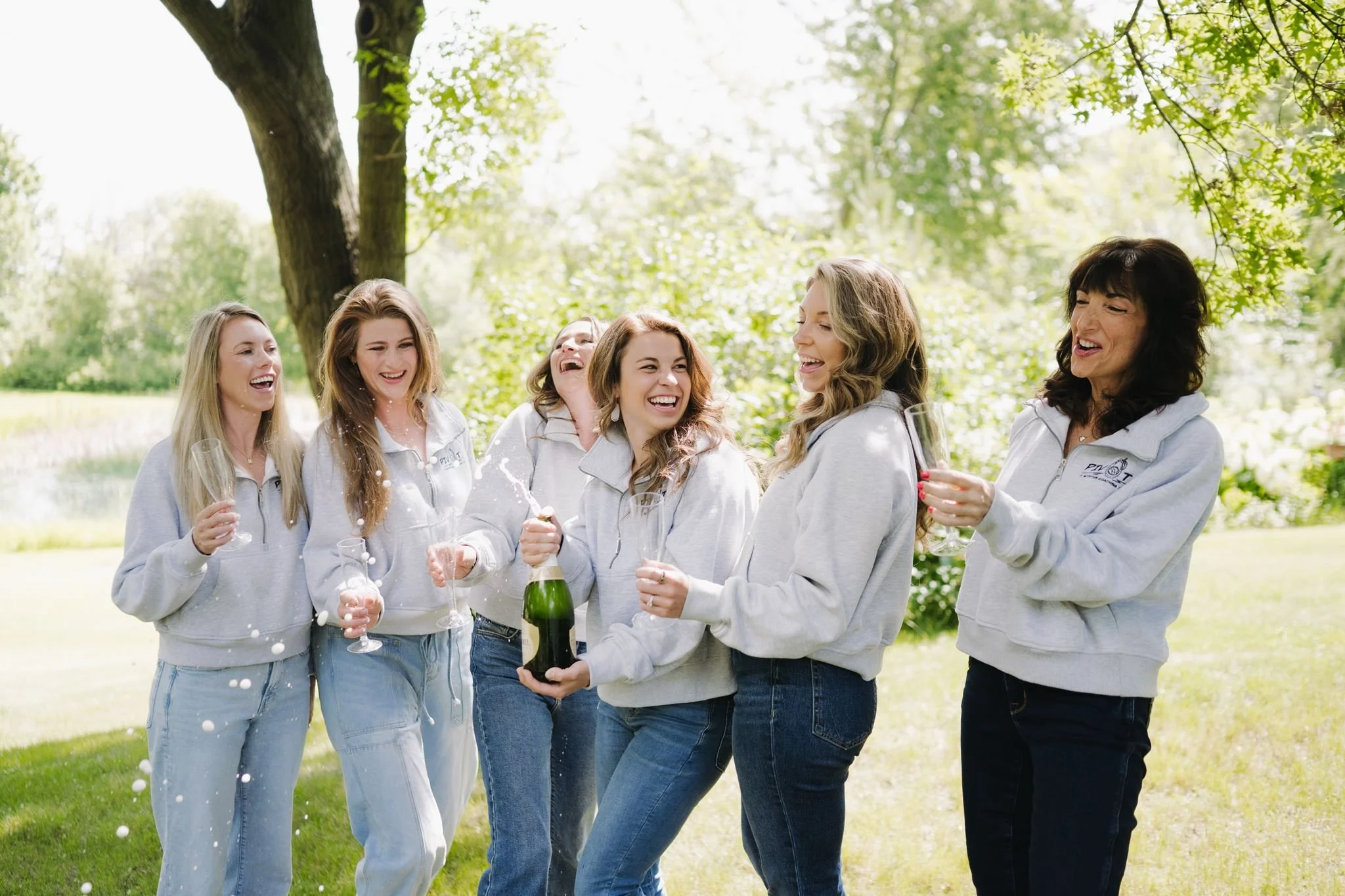 Six Pivot dietitians located in grand forks, nd celebrating outdoors with glasses of champagne, some wearing matching gray hoodies, under a large tree on a sunny day