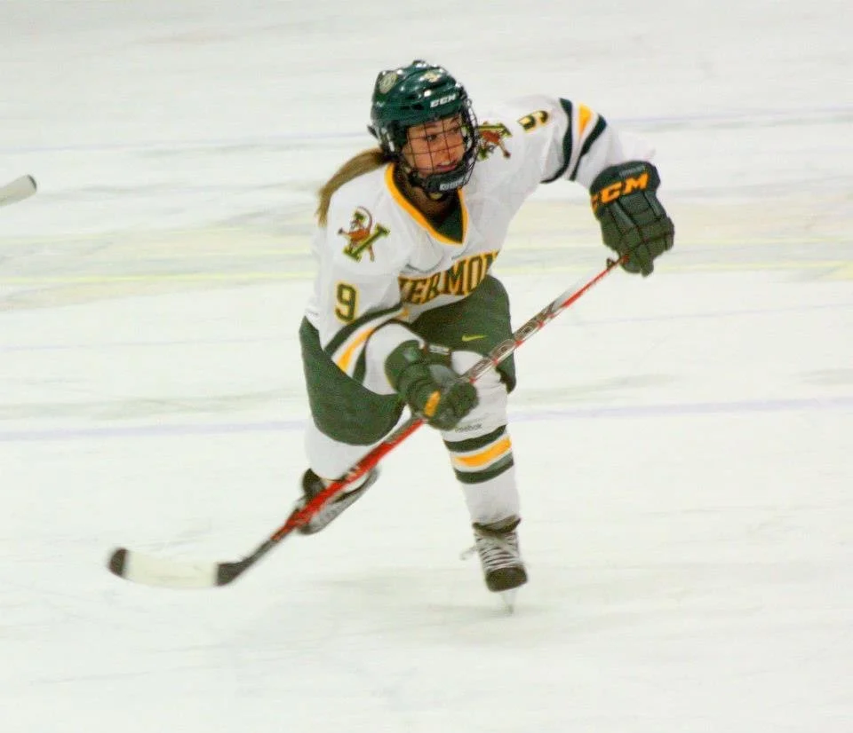 Certified Sports Dietitian female hockey player skating on ice, wearing a white jersey with black, yellow, and green details, black helmet, and black gloves, holding a hockey stick.