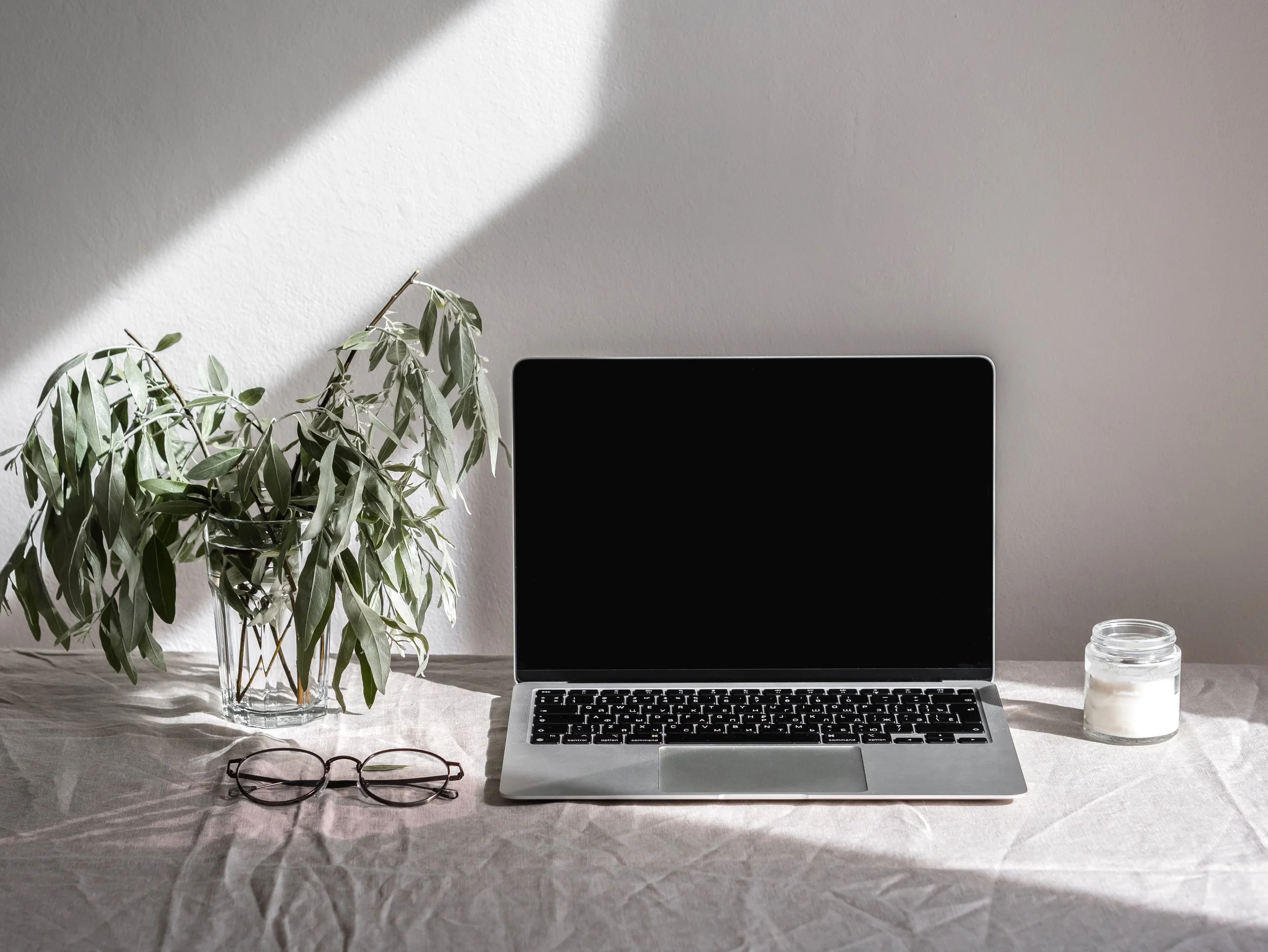 A workspace with a laptop, glass of water or candle, a pair of eyeglasses, and a potted plant on a textured beige table against a plain wall. Sunlight casts shadows on the wall.