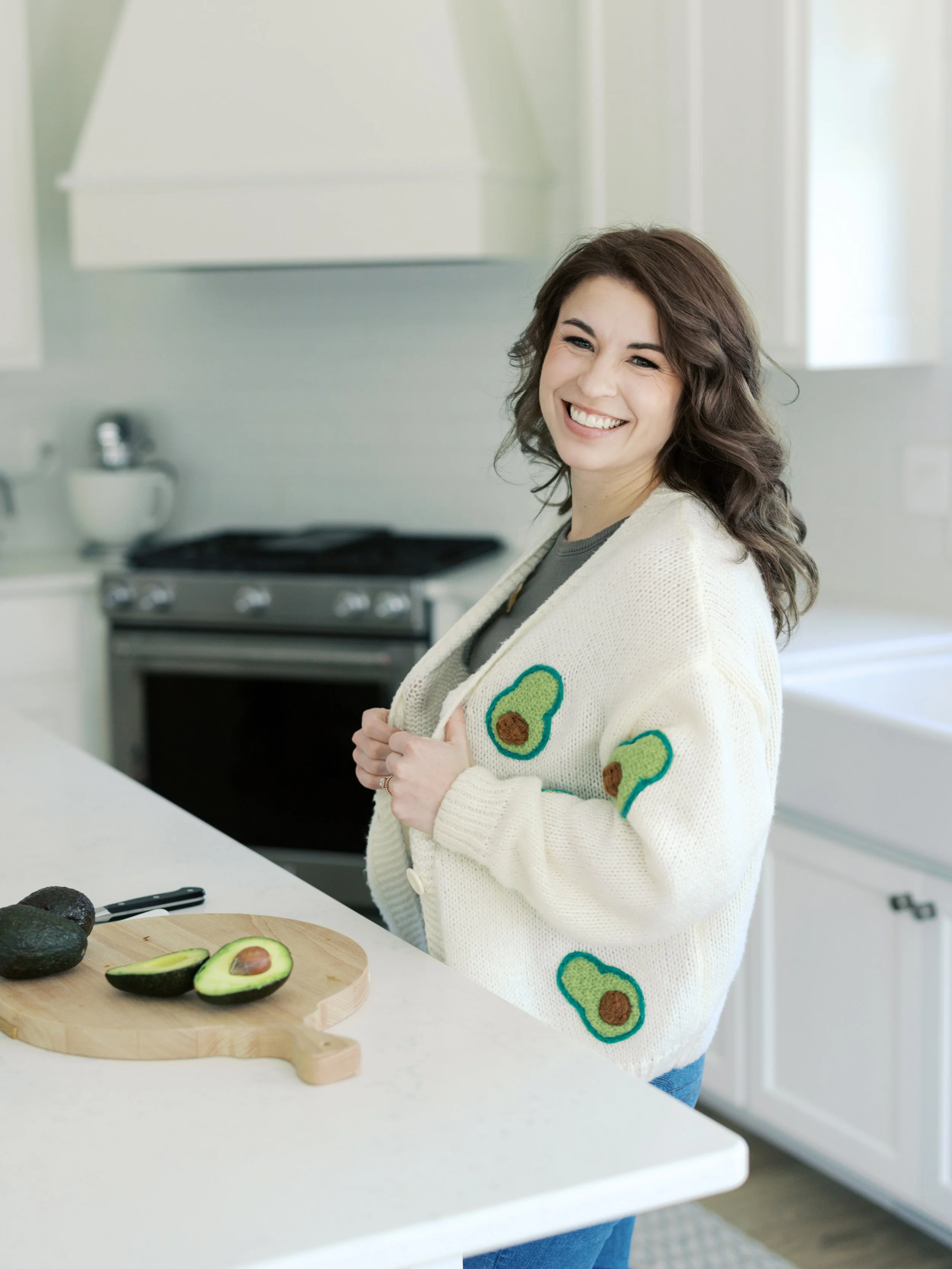 Registered dietitian and nutritionist serving Grand Forks, ND smiling in a kitchen, standing near a white counter with chopped avocado and whole avocados on a wooden cutting board, wearing a cream sweater with avocado designs.