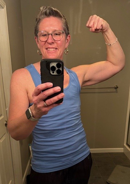 Woman in a blue tank top and glasses flexing her right arm muscle in a bathroom mirror.