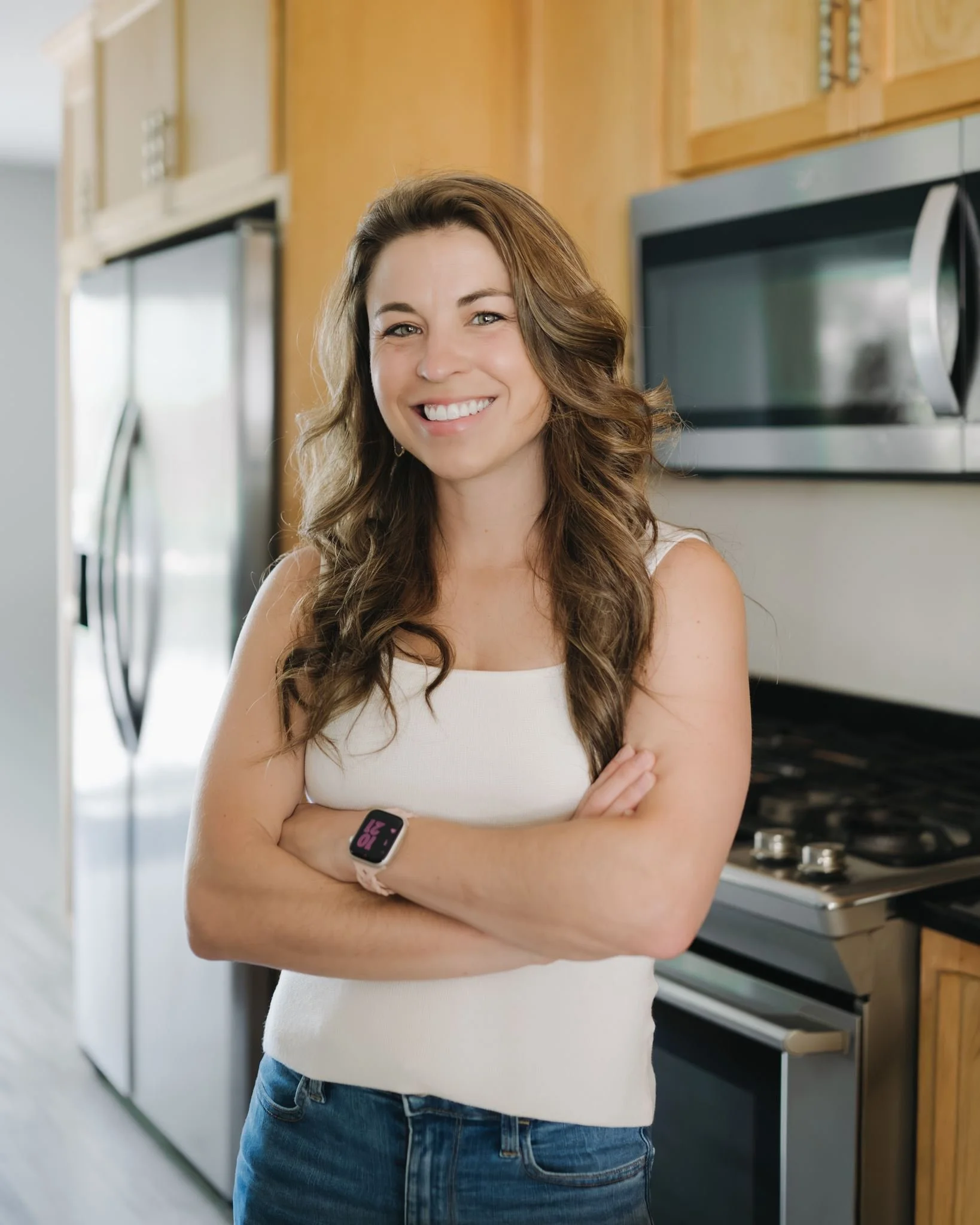 A sports nutritionist standing in a kitchen, smiling, with her arms crossed, wearing a white tank top, blue jeans, and a smartwatch on her left wrist.