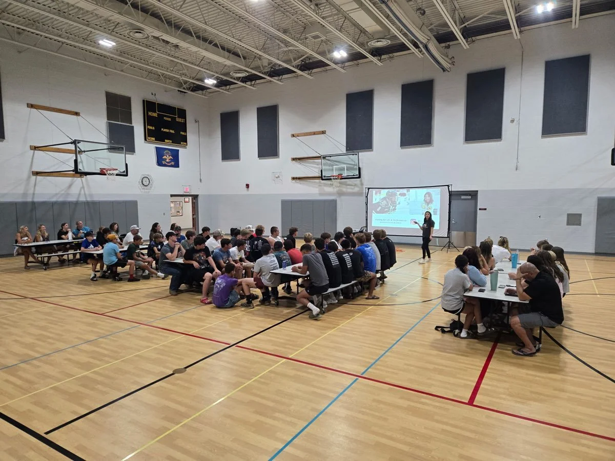 A classroom or gymnasium with a presentation being given to students seated at tables. The keynote wellness speaker stands near a large projection screen on the right side of the room.