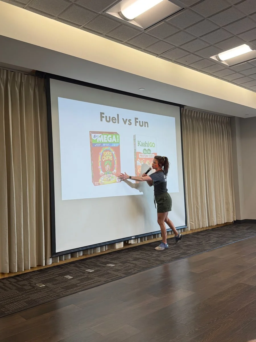 A sports dietitian giving a presentation in front of a projected slide titled 'Fuel vs Fun,' comparing cereal products. She is holding a microphone and gesturing toward the slide, which displays images of cereal boxes.