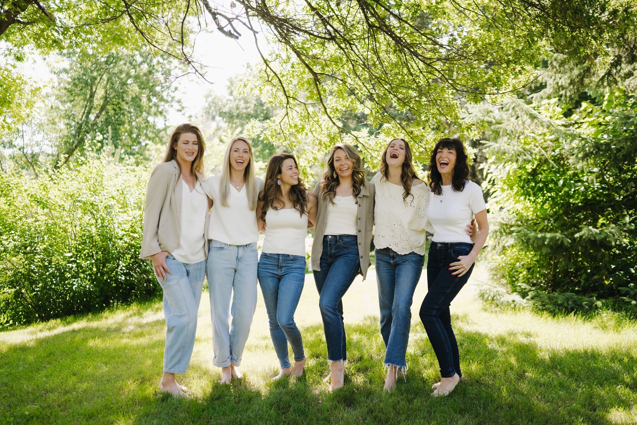 Group of six Pivot nutrition coaches standing together outdoors on grass under a large leafy tree, smiling and laughing