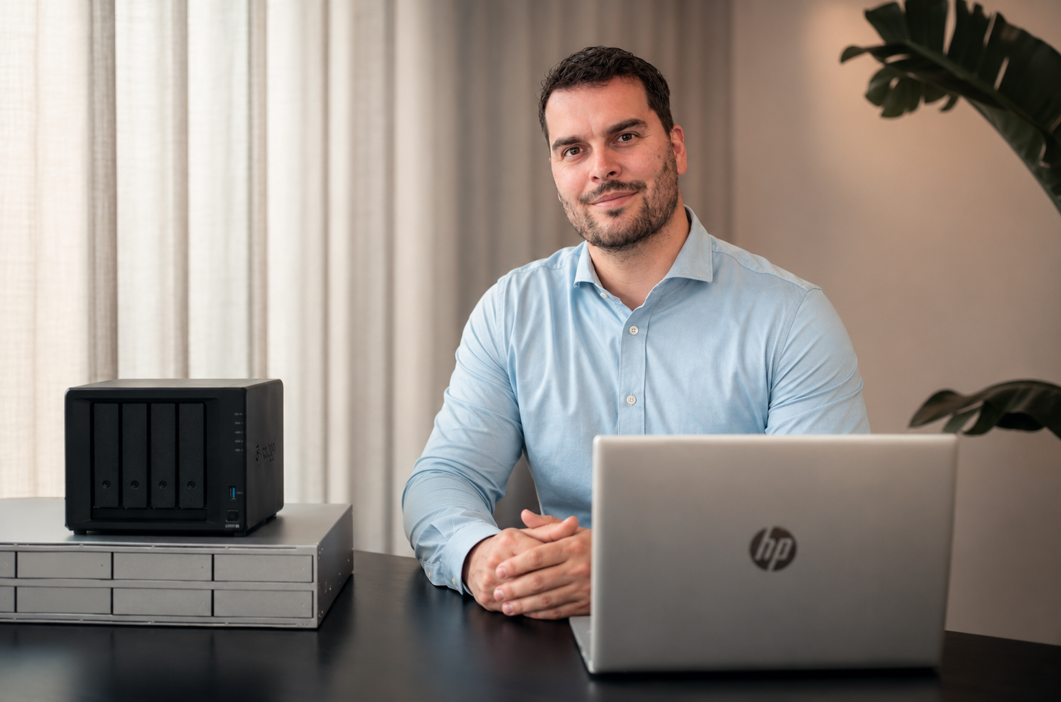 Man zit aan een bureau met een HP-laptop, een NAS-server en een plantenblad op de achtergrond.