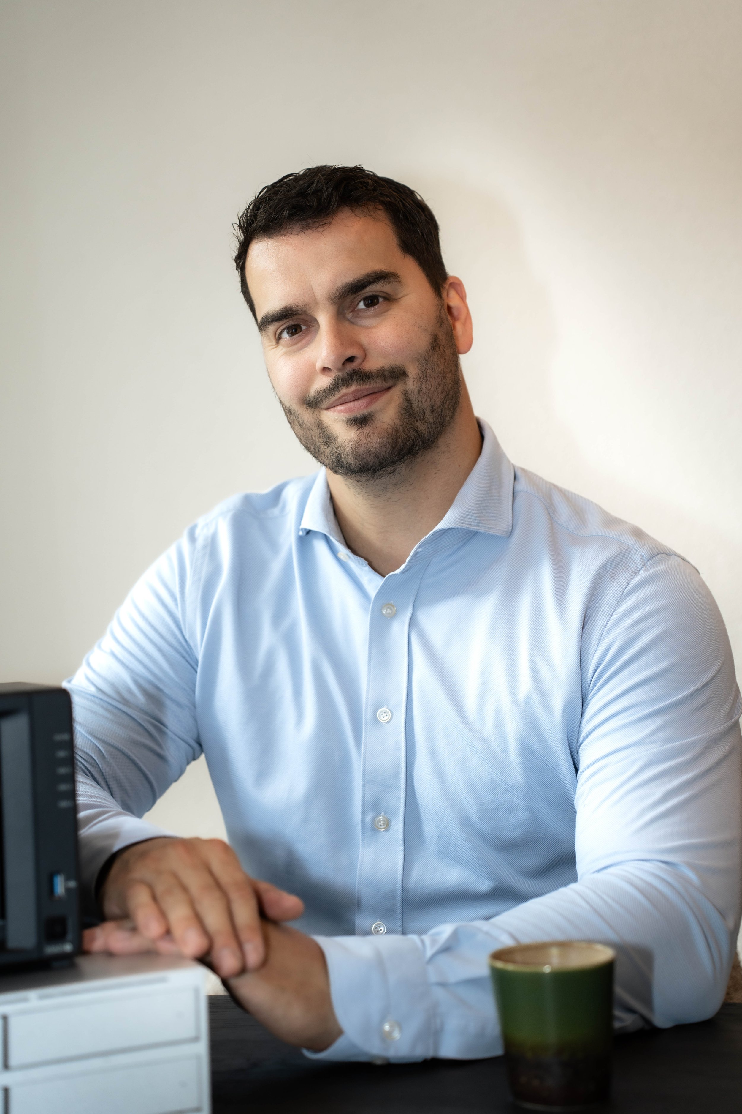Portret van een man met donker haar en een lichte baard, gekleed in een lichtblauw overhemd, zittend aan een bureau, met zijn handen gekruist. Op de voorgrond zitten een kop koffie en een apparaat.