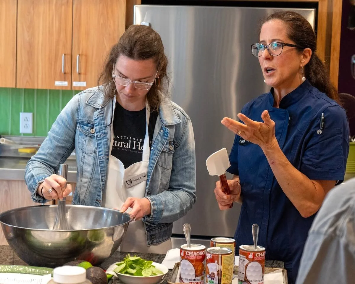 Two women are in a kitchen setting, one is stirring a large metal mixing bowl, and the other is talking and gesturing with her hand. There are cans of Chaokoh coconut milk and a plate of leafy greens on the counter.