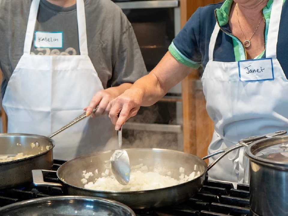 Two women cooking on a stovetop, one stirring diced onions in a pan and the other preparing to add ingredients, both wearing aprons and name tags that read 'Katelin' and 'Janet'.