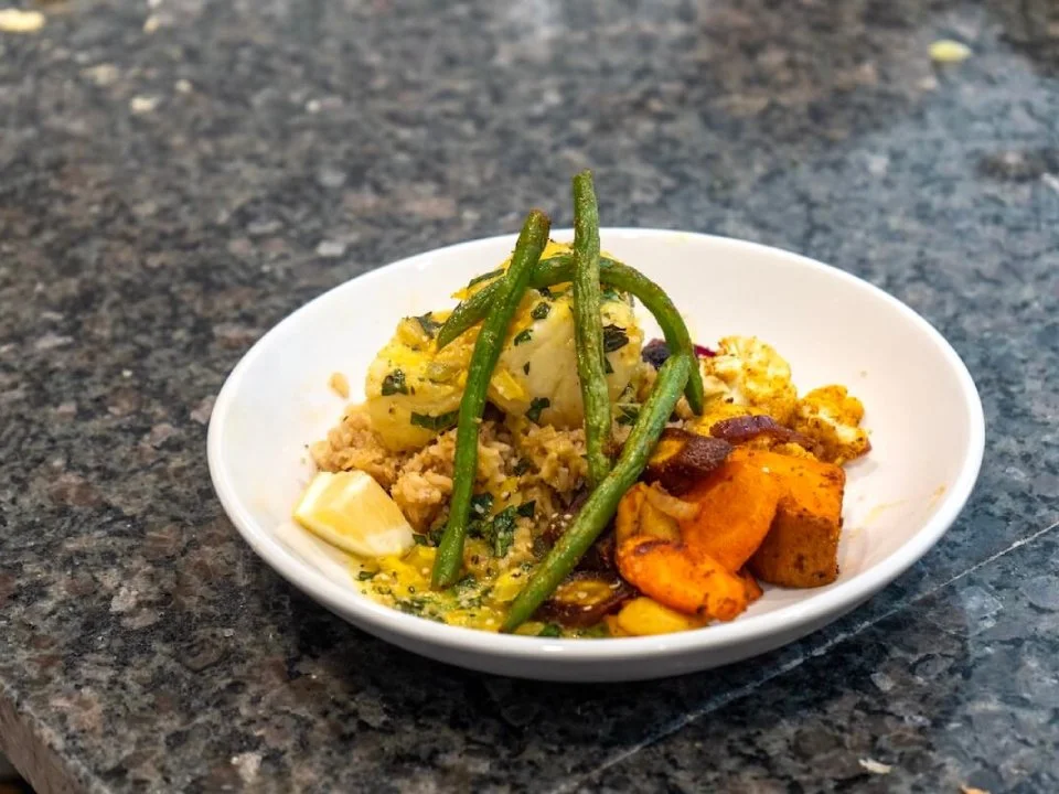 A small white bowl of Indian curry with chunks of vegetables and spices, topped with green beans and accompanied by a lemon wedge, placed on a dark granite countertop.