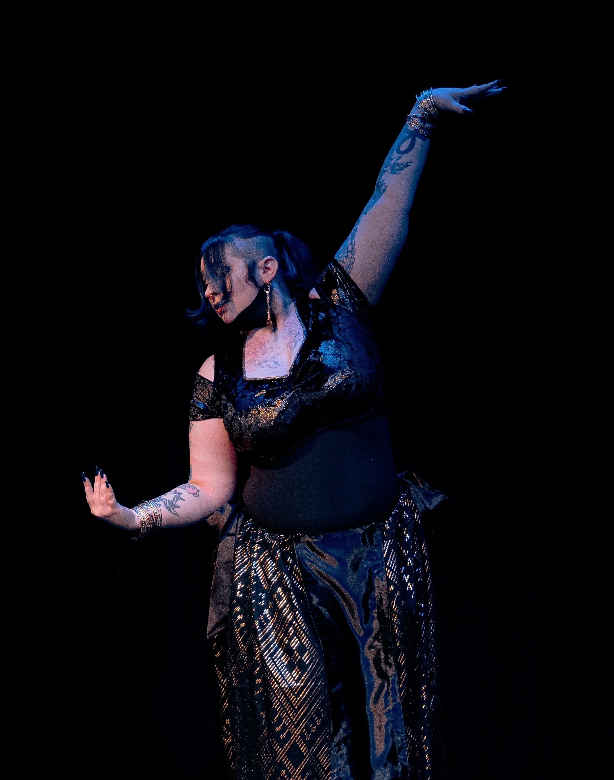 A woman dancing on stage with her arm raised, wearing a black and gold outfit, with dark hair and tattoos on her arm, illuminated by stage lighting against a black background.