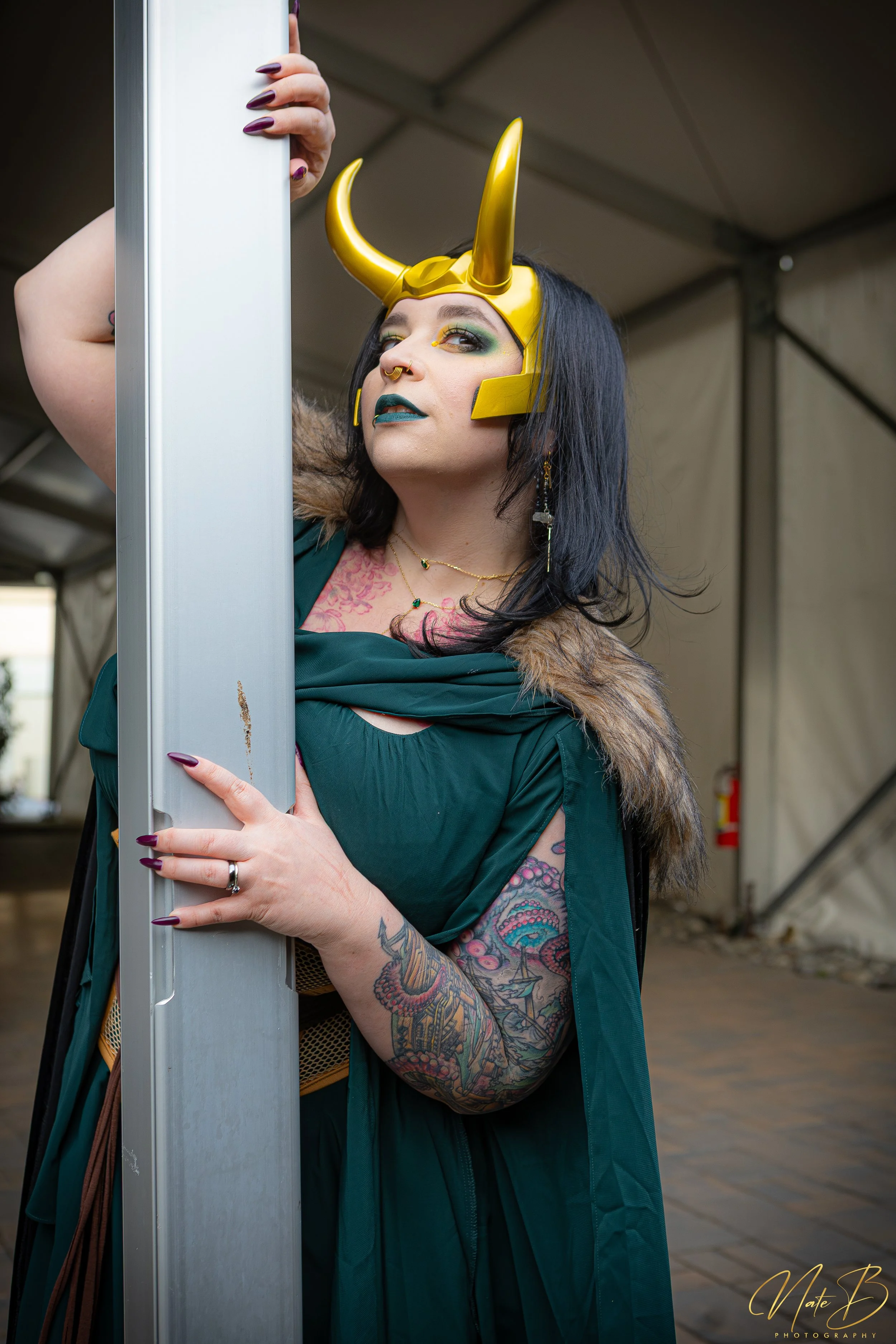 Person with tattoos, dark hair, and bold makeup wearing a gold Viking helmet with horns, taking a selfie or photo with a cellphone, inside a tent or large portable shelter.