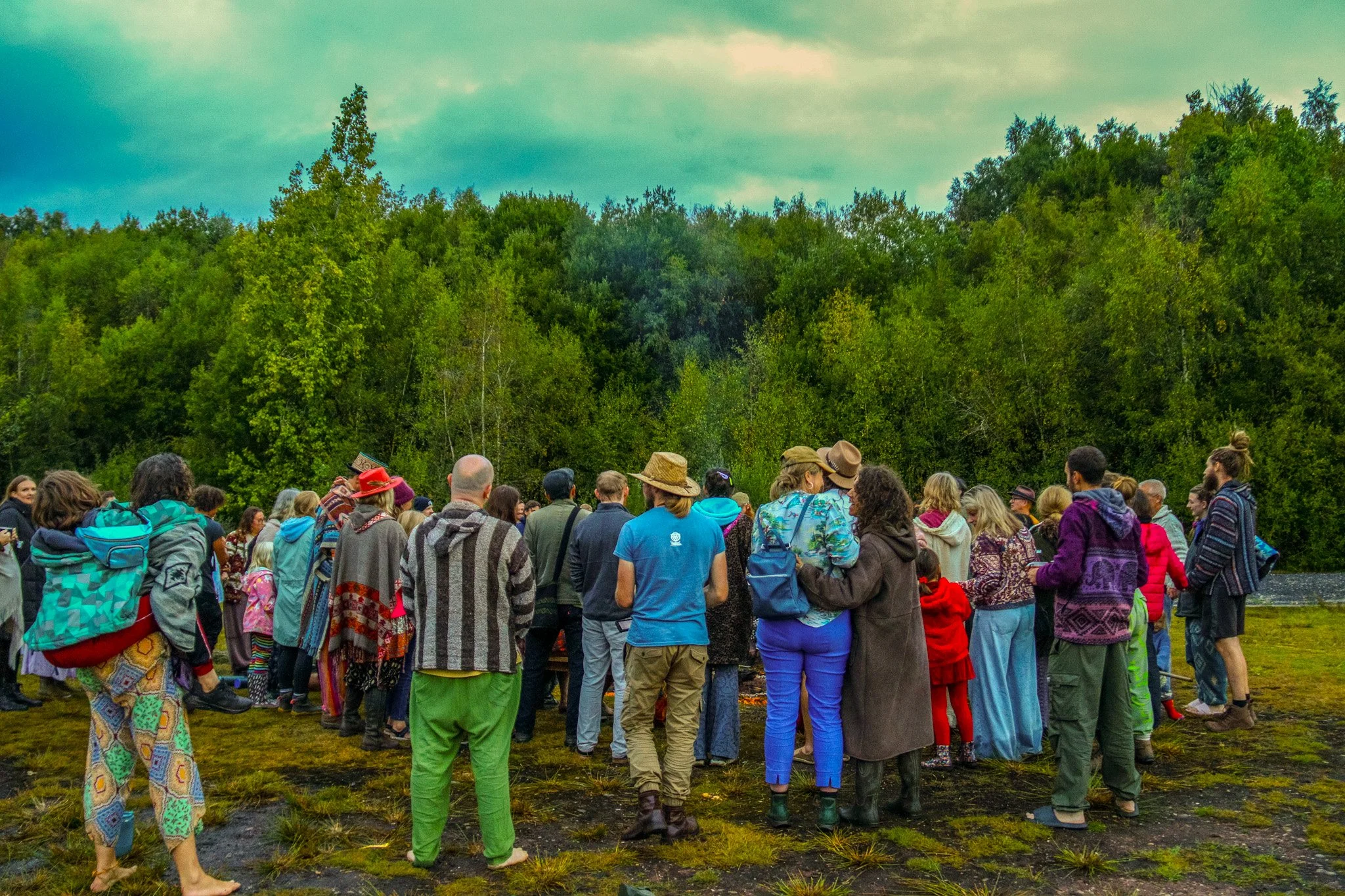 A large group of diverse people gathered outdoors on a grassy field in front of a dense forest, some wearing colorful and patterned clothing, with many backpacks and hats, under an overcast sky.