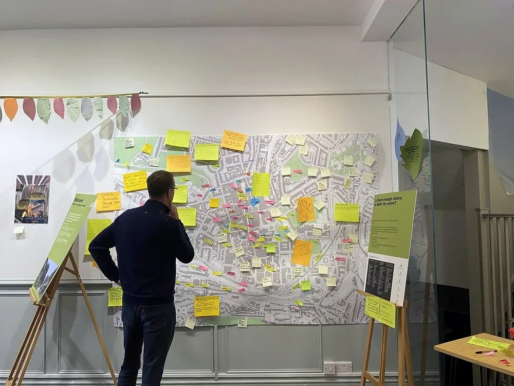 A man is studying a large map on a wall covered with colourful sticky notes, in a room with informational display boards and a glass partition.
